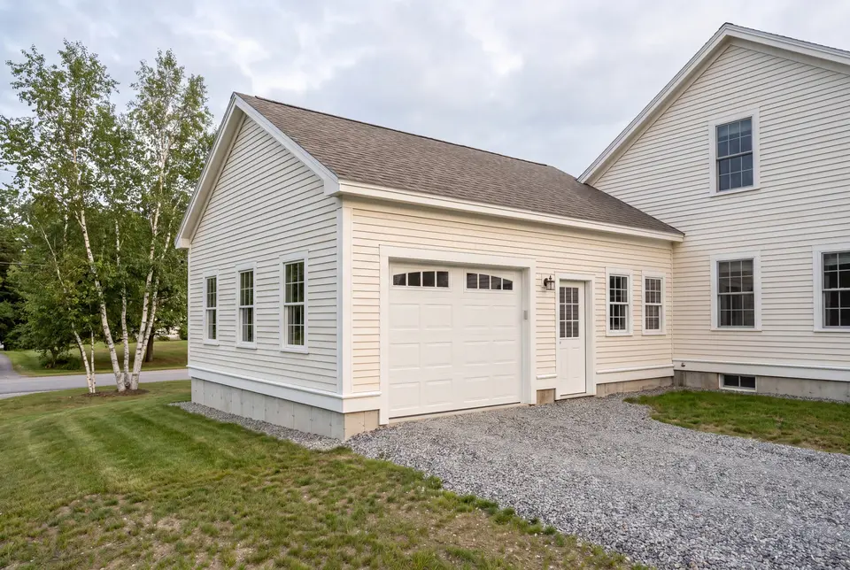garage builder in Westbrook, ME: illustrative scene, A newly built, single-bay garage with white siding and a brown roof, attached to a house, with a gravel driveway and surrounding green grass