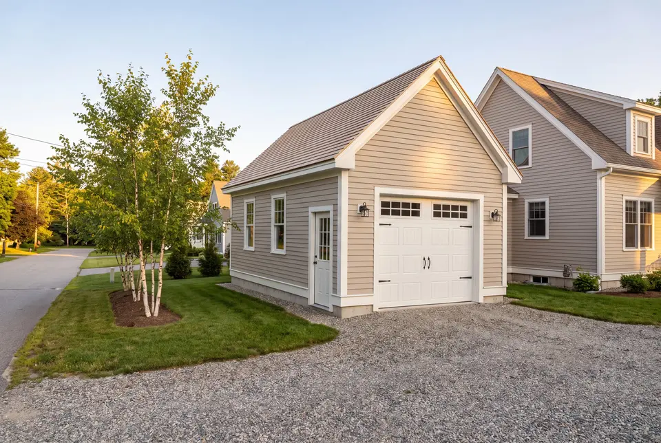 Illustration: A newly built, light-colored detached garage with a gravel driveway next to a house, surrounded by green grass and trees