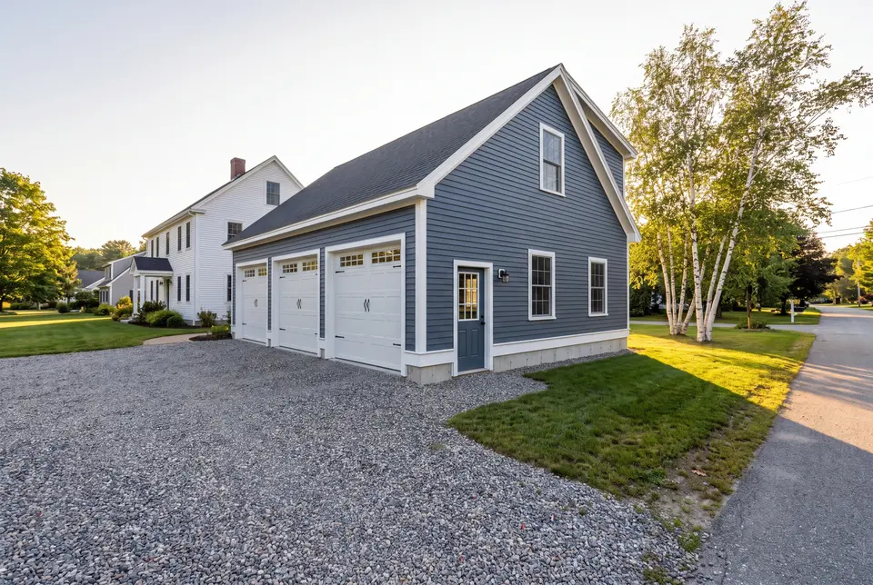 Illustration: A newly built, detached three-bay garage with deep slate blue siding and white trim, next to a white house, with a gravel driveway and green lawn