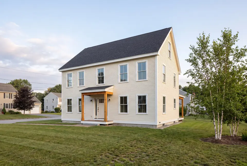 general contractor in Biddeford, ME: illustrative scene, A newly built two-story farmhouse with light yellow siding, a dark gray roof, and a wooden porch, set on a green lawn with a birch tree