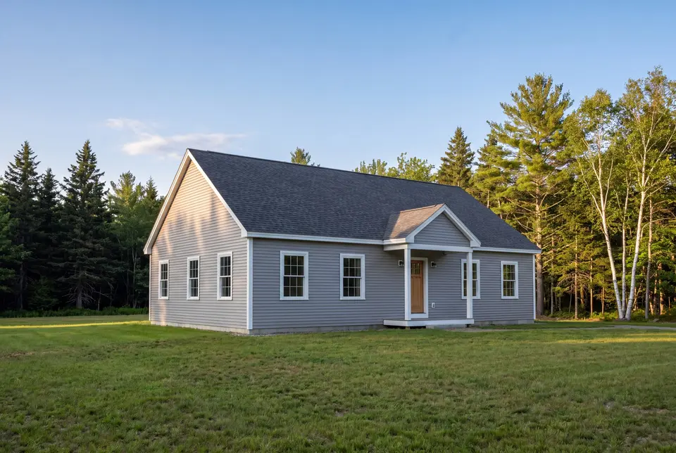 Illustration: A newly built gray ranch house with a dark roof and white trim, surrounded by a green lawn and trees under a blue sky