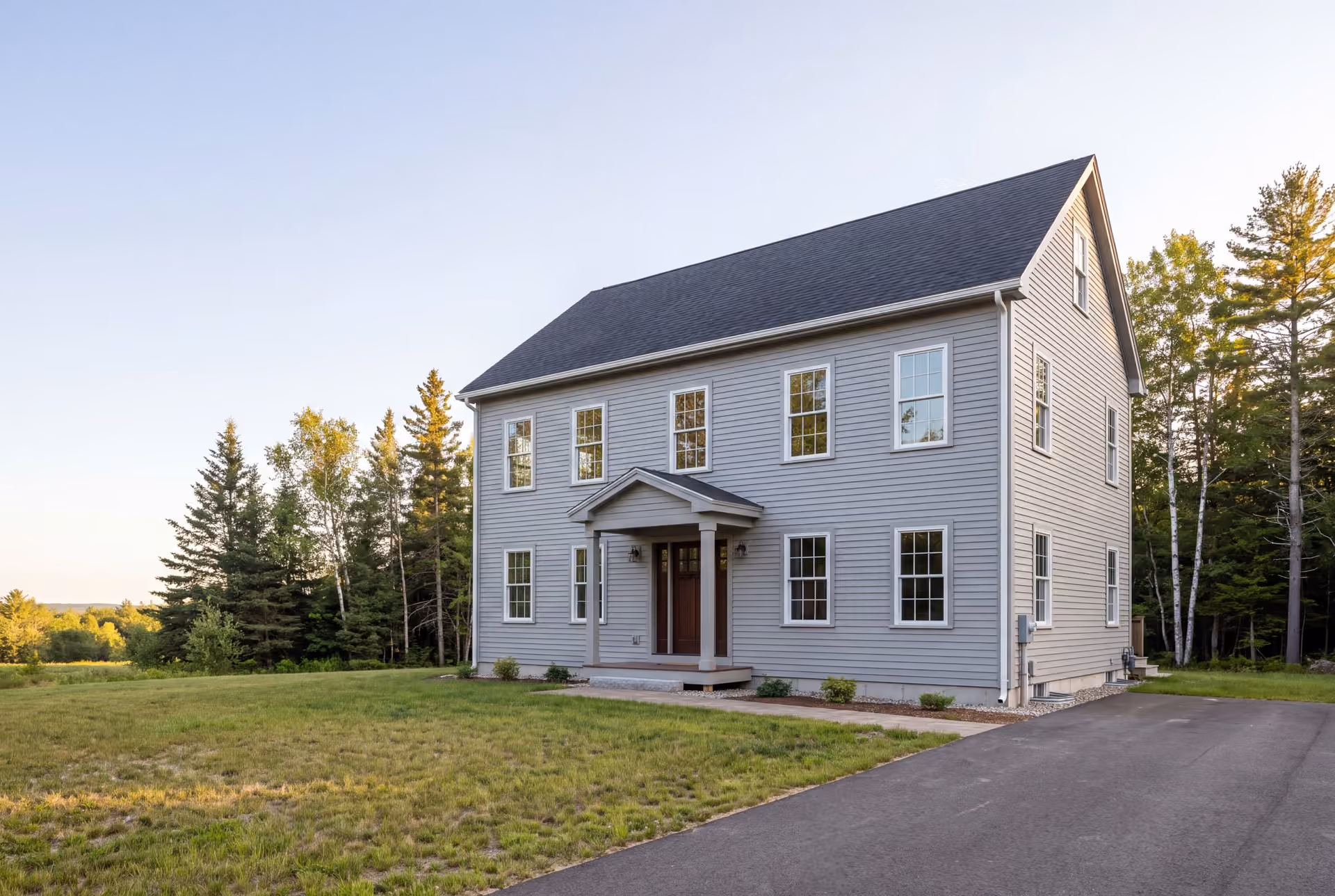 general contractor in Cape Elizabeth, ME: illustrative scene, A newly built two-story Colonial house with gray siding and a dark roof, viewed from the front and side, surrounded by a lawn and trees
