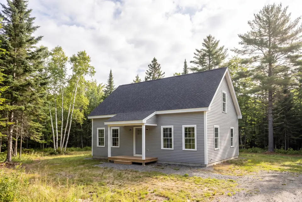 Illustration: A newly built gray farmhouse with a dark roof and white trim, surrounded by trees and a grassy, dirt area