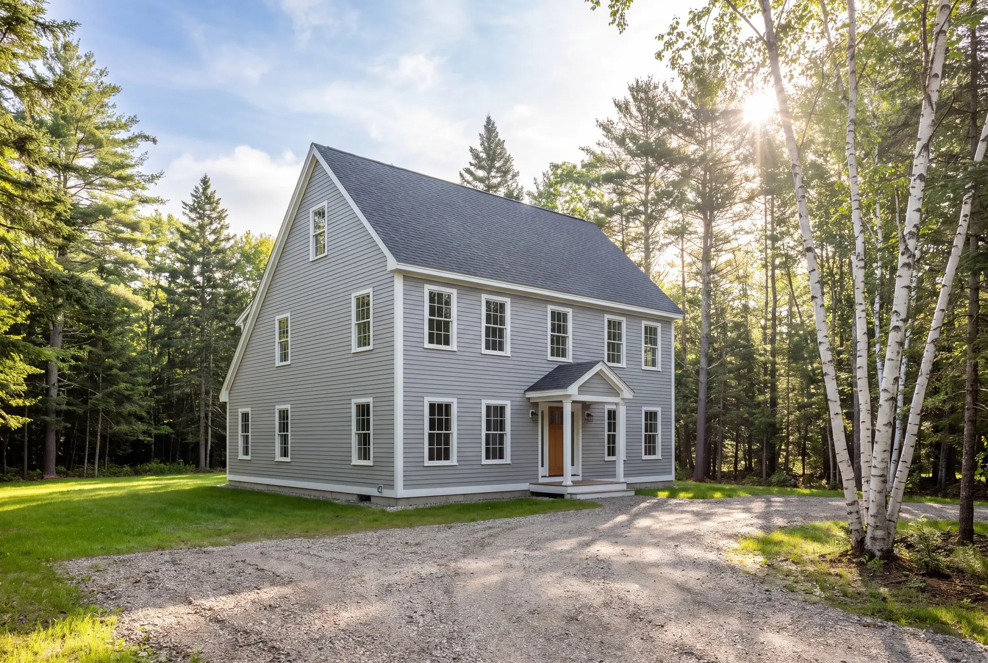 Illustration: A newly built gray saltbox house with white trim and a dark roof, set against a backdrop of green grass, a gravel driveway, and tall trees