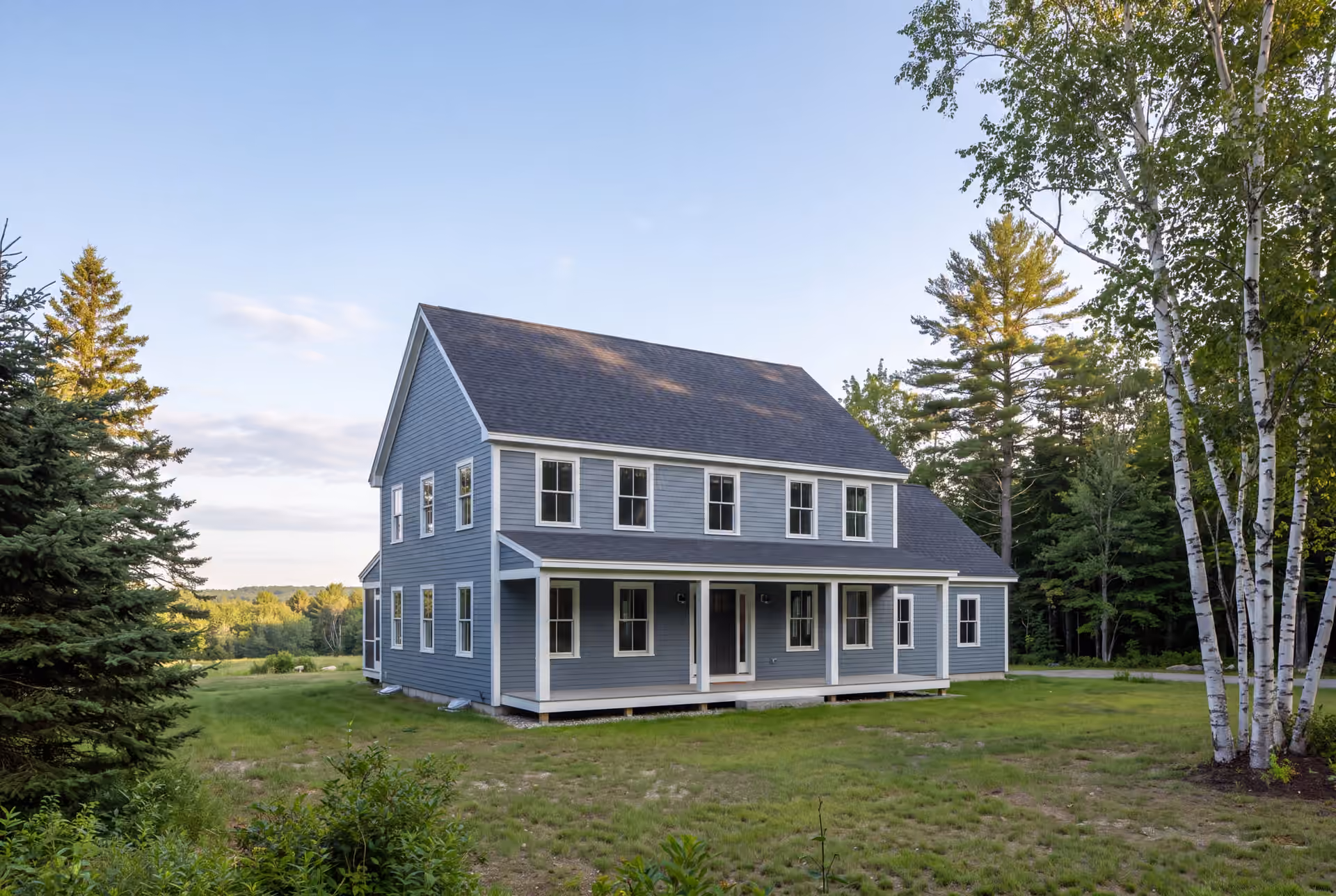 general contractor in Freeport, ME: illustrative scene, A newly built two-story blue-gray house with a dark roof and white trim, featuring a front porch, surrounded by green grass and trees under a clear sky