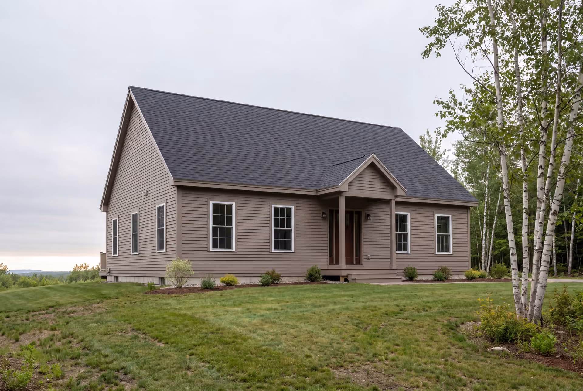 general contractor in Gorham, ME: illustrative scene, A newly built single-story house with taupe siding and a dark roof, viewed from a three-quarter angle, showing the front and side elevations, surrounded by grass and trees