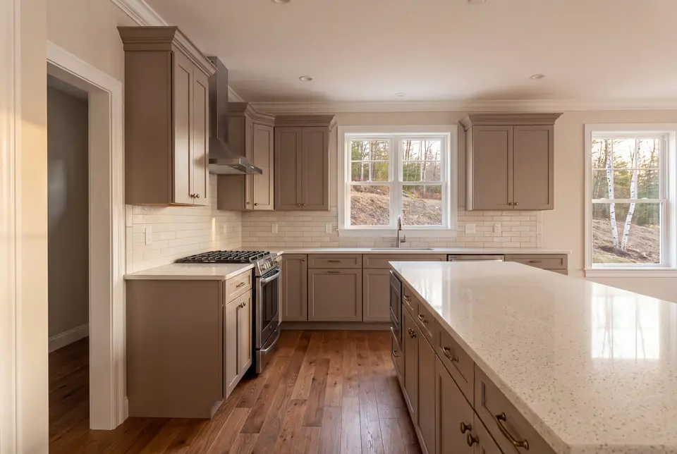 Illustration: A newly finished kitchen with taupe shaker cabinets, a quartz island, oak flooring, and white-trimmed windows looking out to a wooded area