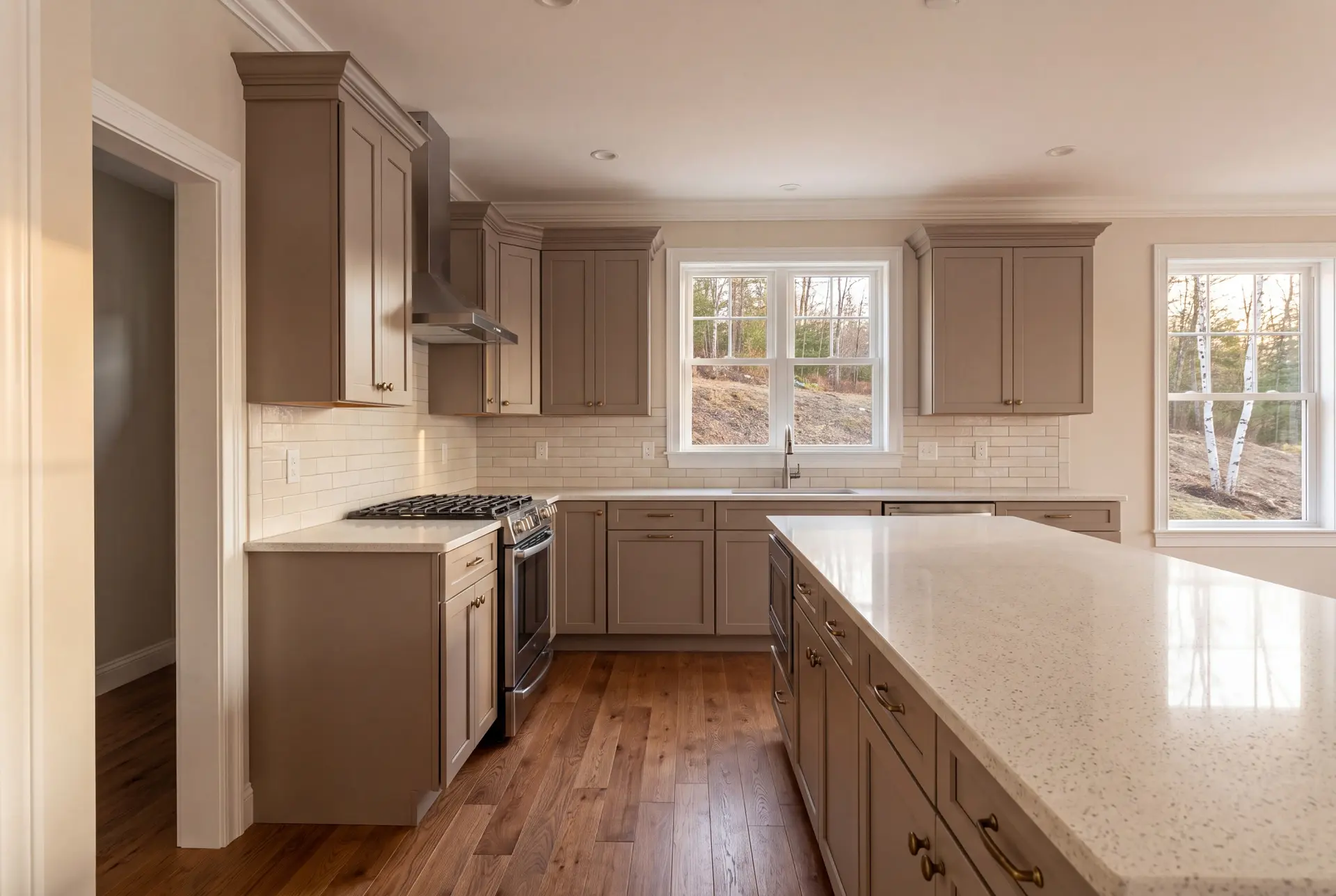 Illustration: A newly finished kitchen with taupe shaker cabinets, a quartz island, oak flooring, and white-trimmed windows looking out to a wooded area