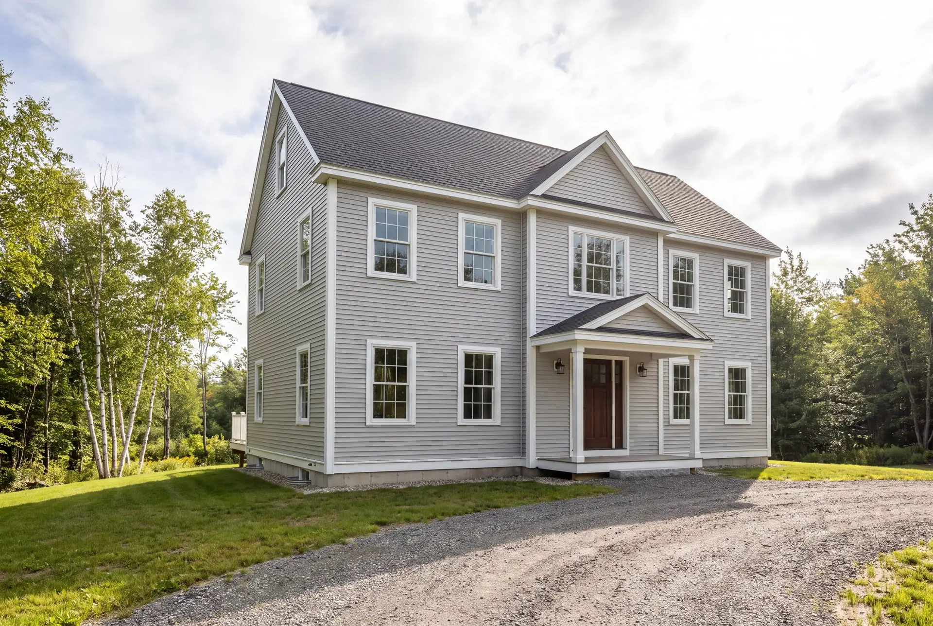 Illustration: A newly built two-story Colonial house with light gray siding and a dark roof, viewed from the front and side, surrounded by a gravel driveway, grass, and trees