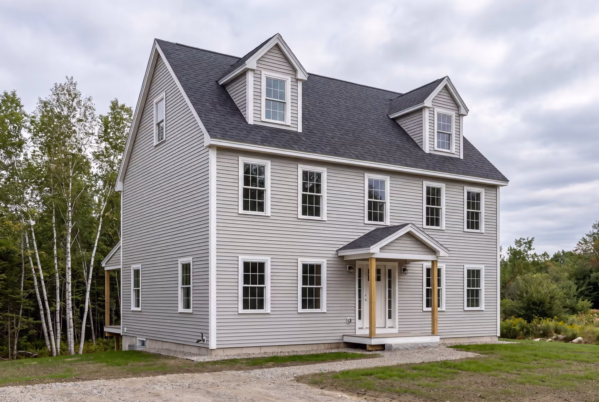 general contractor in Hollis, ME: illustrative scene, A newly built gray Cape-style house with a dark roof and white-trimmed windows, set against a cloudy sky and surrounded by sparse grass and trees