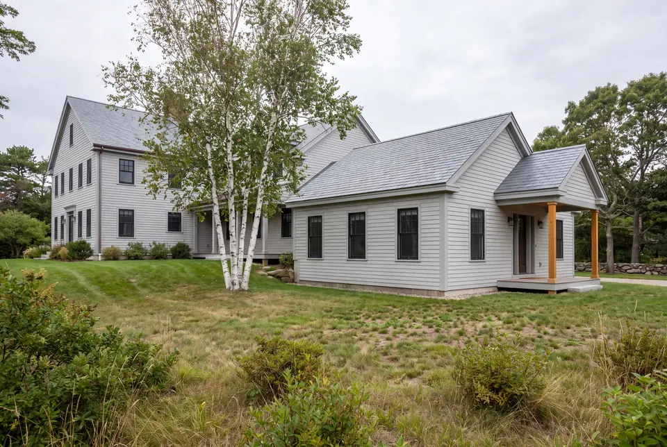 Illustration: A light gray clapboard house with a new single-story addition, featuring matching siding, roof, and windows, set in a grassy landscape with a birch tree