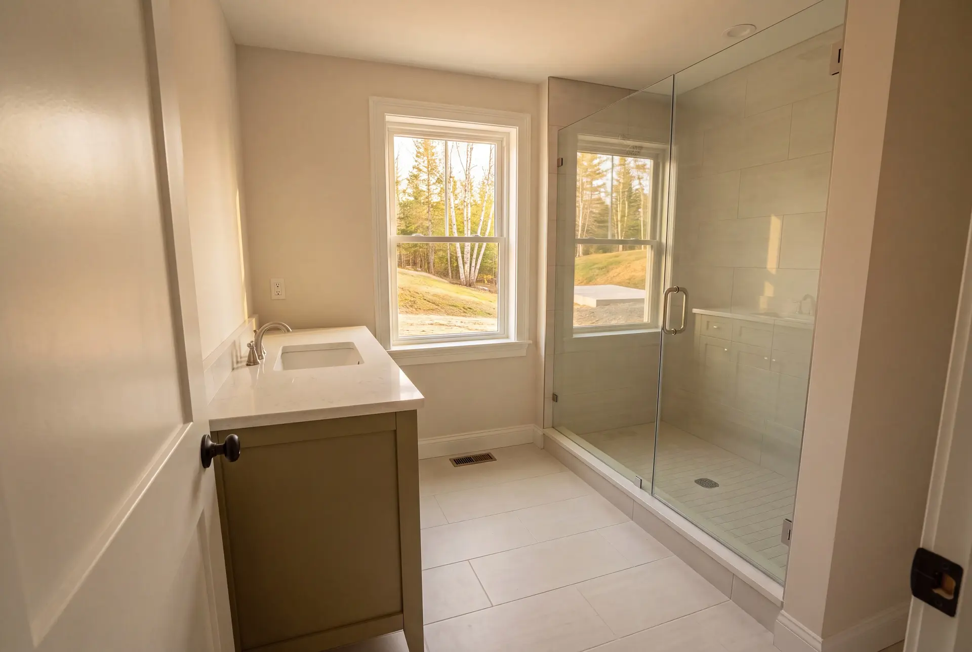 Illustration: A newly finished bathroom featuring a dusty olive wood vanity with a quartz top, a glass shower enclosure, and light-colored porcelain tile throughout. A window overlooks a natural outdoor scene