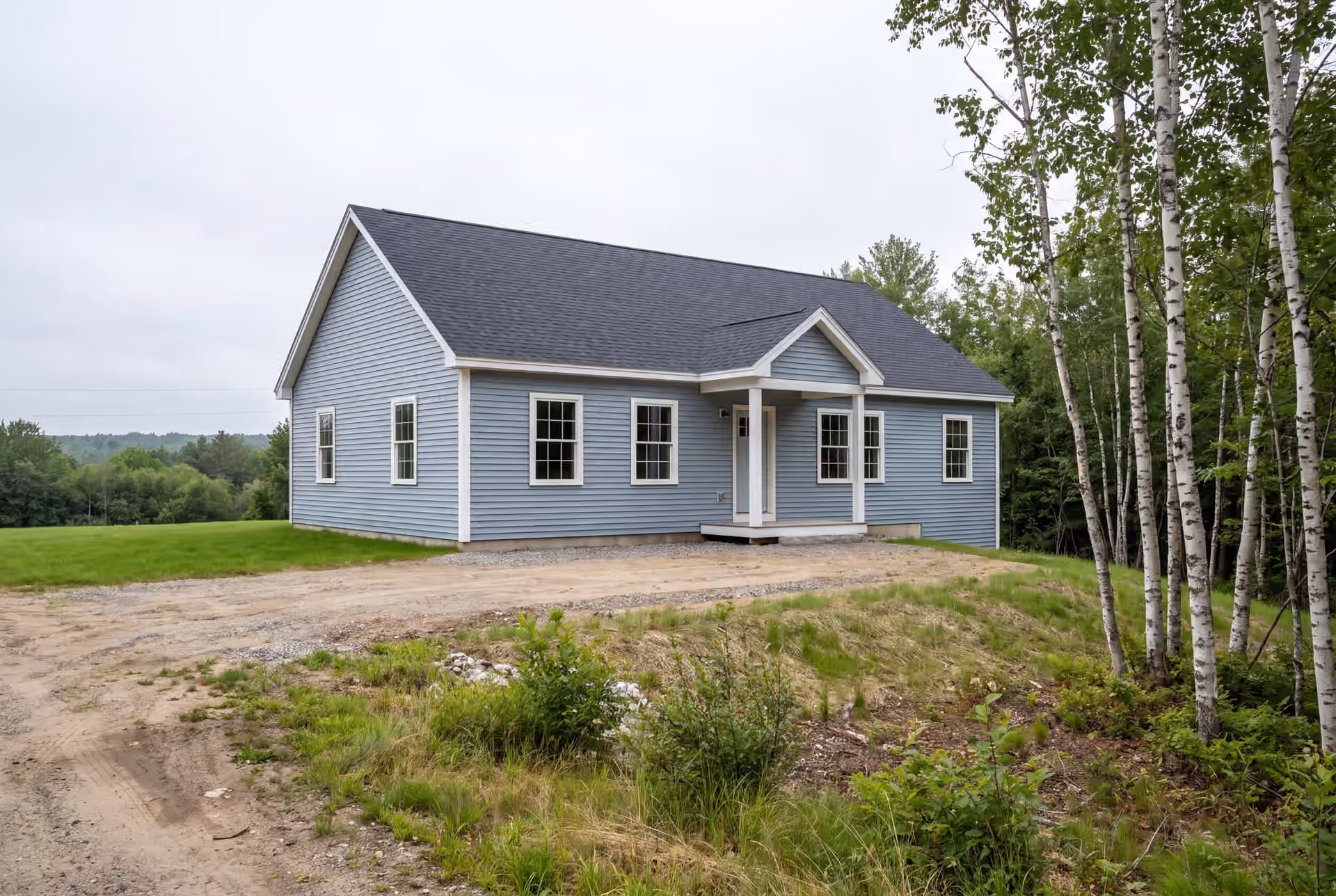 general contractor in Lyman, ME: illustrative scene, A newly built light blue ranch house with a dark roof and white trim, set on a partially cleared lot with trees