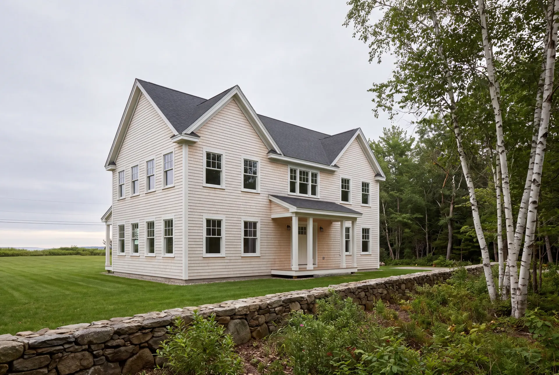 Illustration: A newly built two-story shingle-style house with light-colored siding and a dark roof, surrounded by a green lawn and a stone wall, with trees on the right