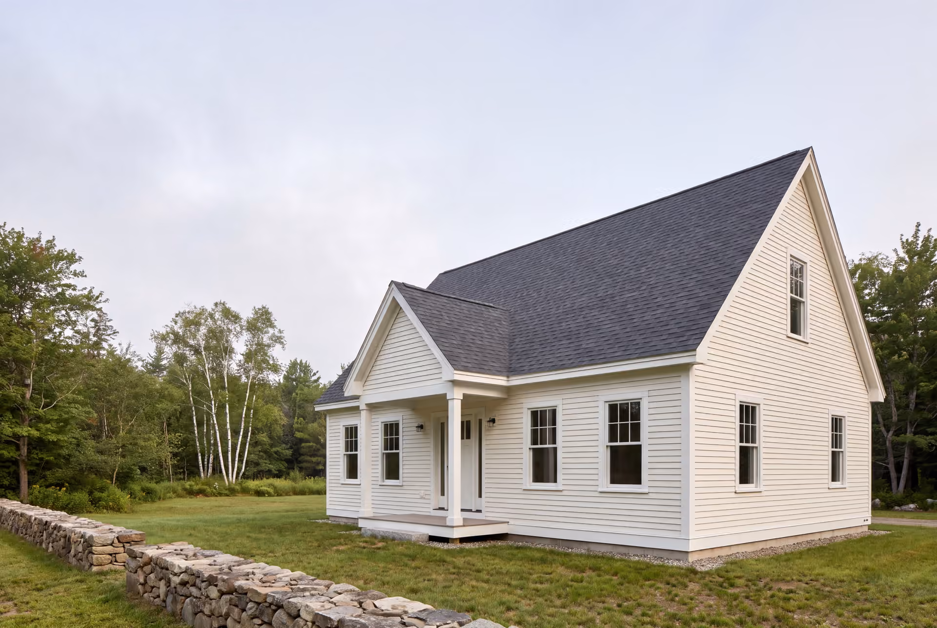 general contractor in Saco, ME: illustrative scene, A newly built white clapboard cottage with a dark gray roof and a covered porch, viewed from a three-quarter angle, surrounded by green grass and trees