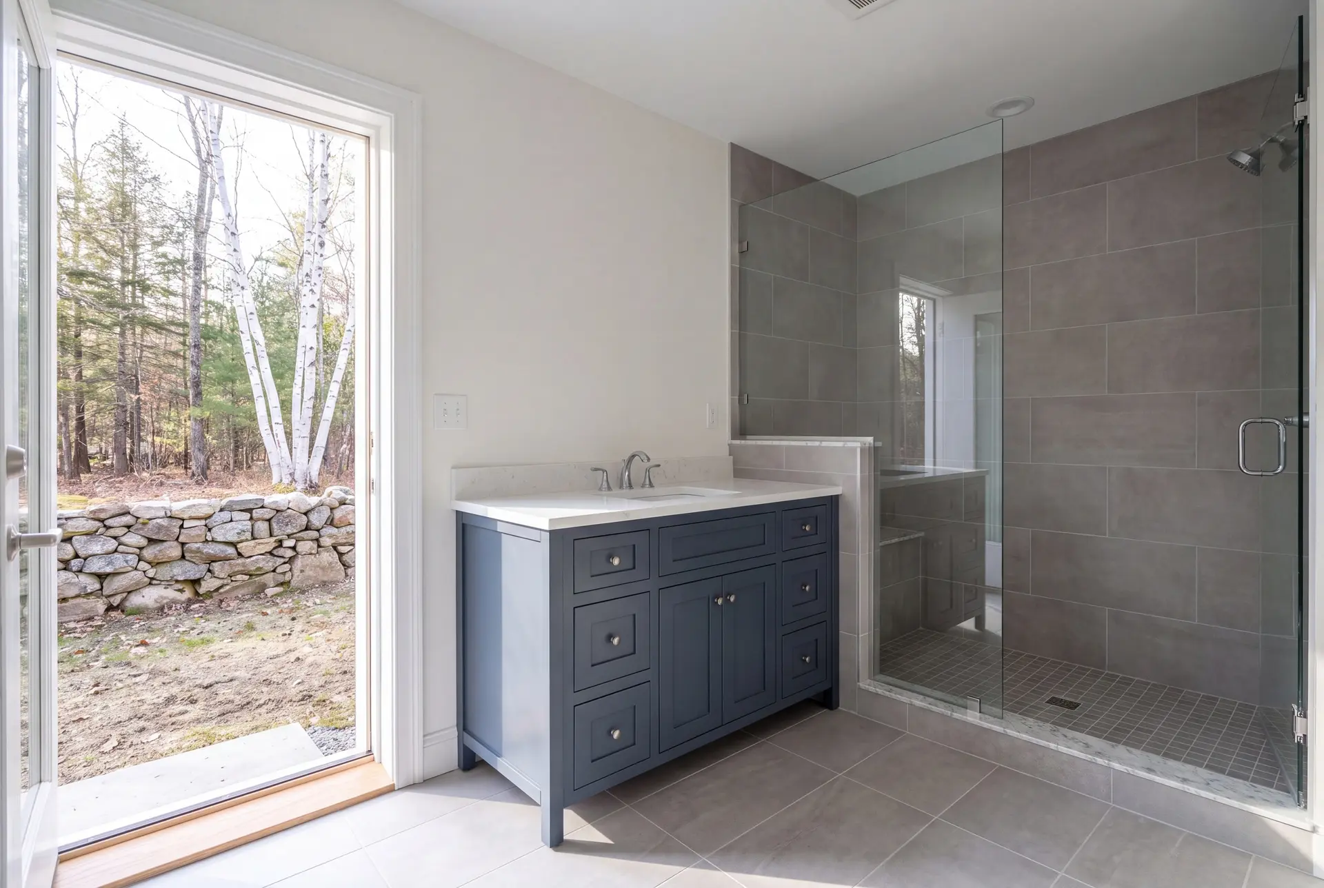 Illustration: A newly finished bathroom featuring a deep slate blue vanity with a white quartz top, a glass shower, and gray tiled floors and shower walls. An open door reveals a natural outdoor scene