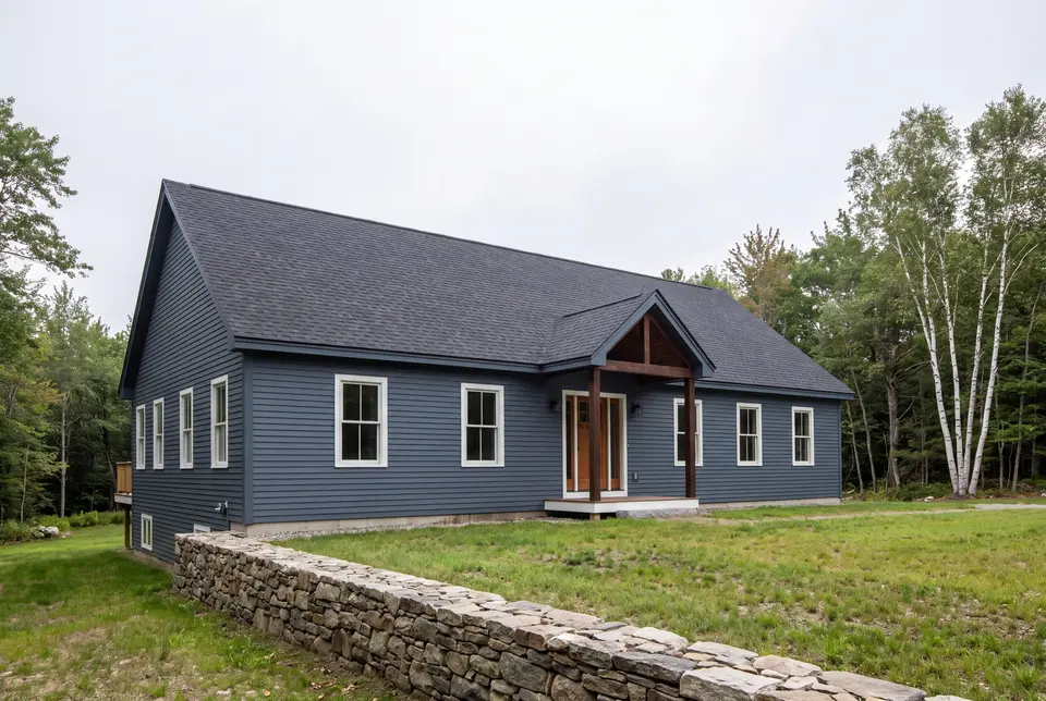 general contractor in Scarborough, ME: illustrative scene, A newly built single-story house with blue siding, a dark roof, and a covered wooden entry, set in a grassy area with trees and a stone wall