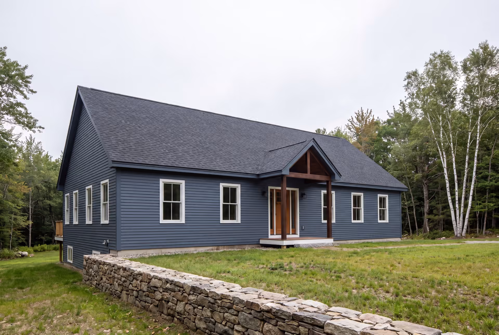 general contractor in Scarborough, ME: illustrative scene, A newly built single-story house with blue siding, a dark roof, and a covered wooden entry, set in a grassy area with trees and a stone wall