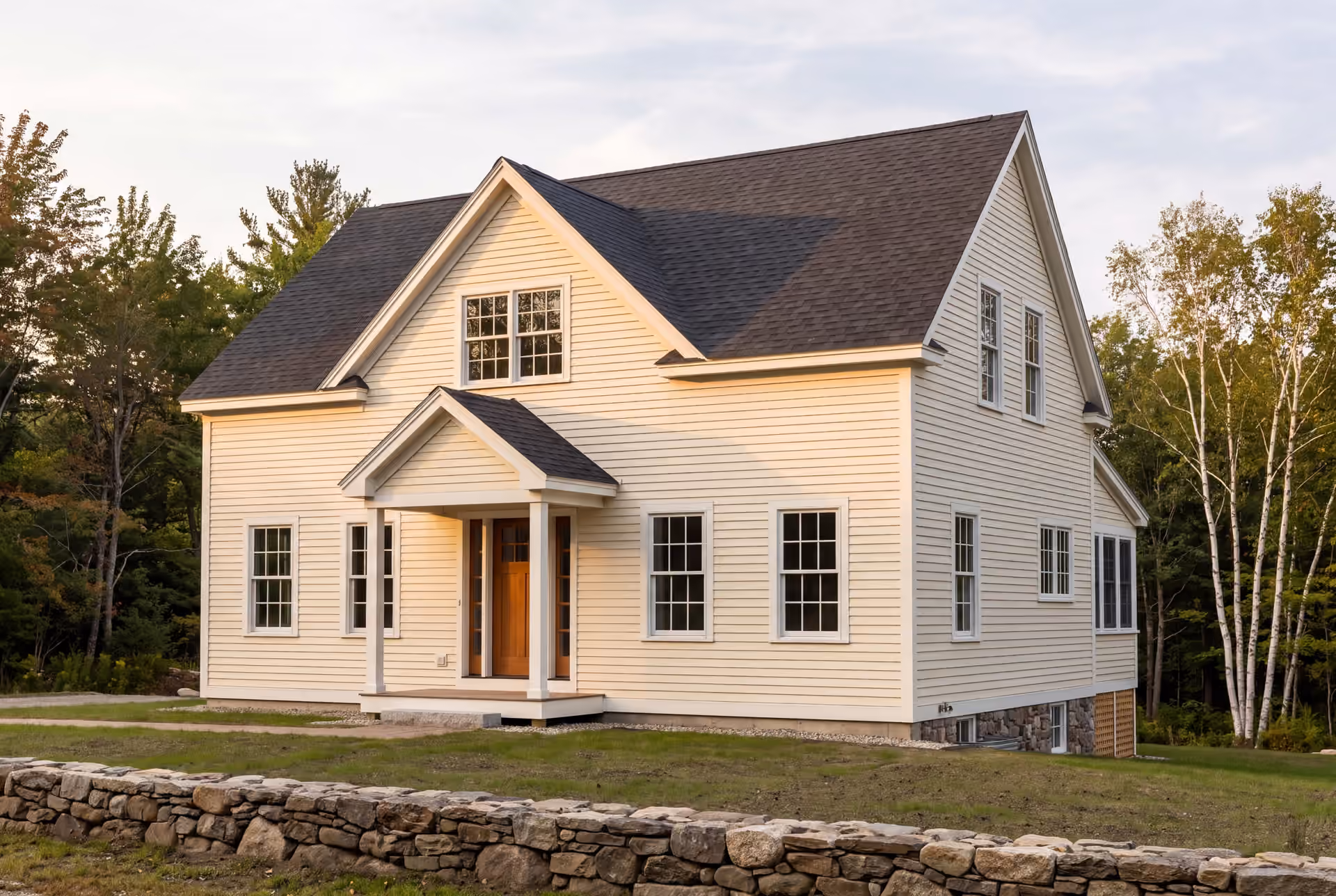 general contractor in South Portland, ME: illustrative scene, A newly built two-story farmhouse with light yellow siding, a dark gray roof, and white-framed windows, seen from a three-quarter angle with a stone wall in the foreground