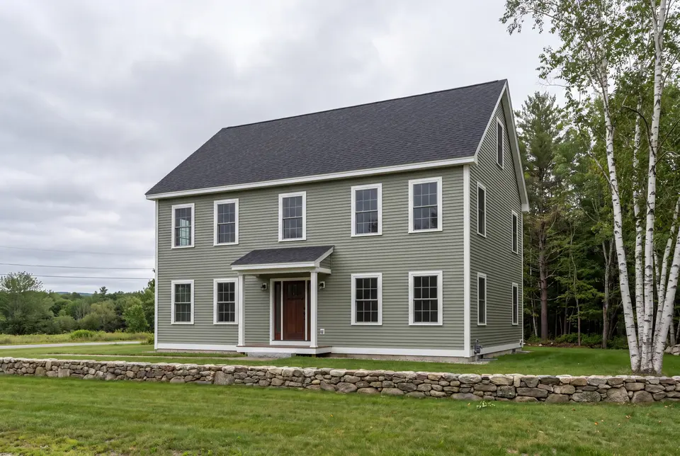 Illustration: A newly built two-story Colonial house with sage green siding and a dark roof, seen from a three-quarter view with a stone wall in the foreground