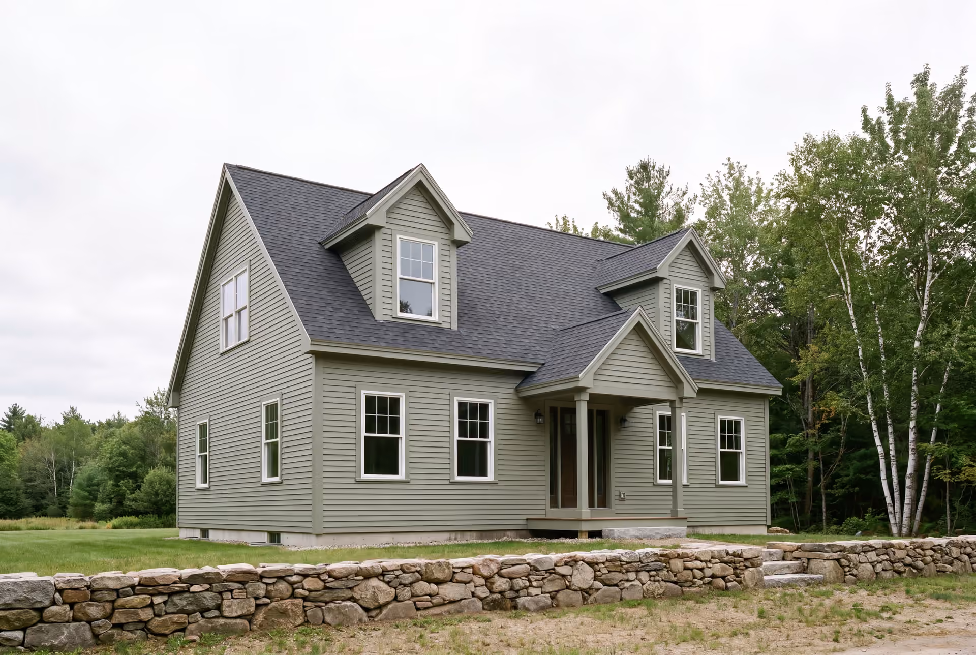 general contractor in Standish, ME: illustrative scene, A newly built Cape house with green siding and a dark roof, viewed from a three-quarter angle, with a stone wall in the foreground