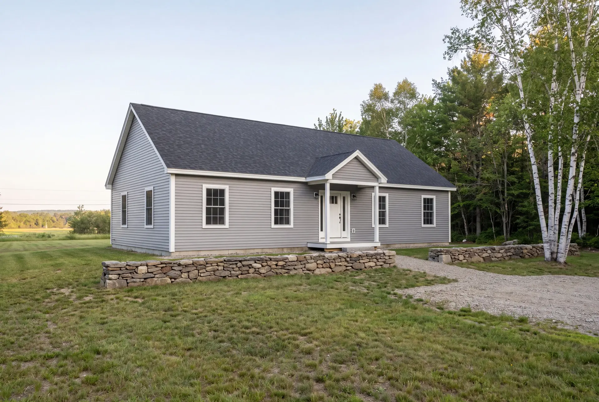 Illustration: A newly built gray ranch house with a dark roof and white trim, seen from a three-quarter view on a grassy lawn with a gravel path and stone wall