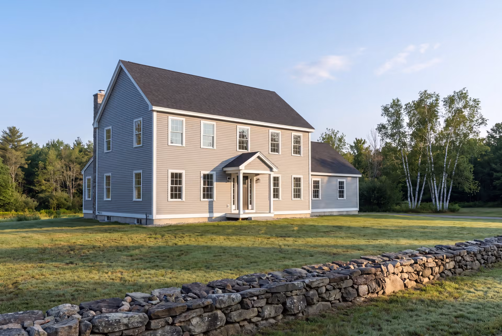 general contractor in Westbrook, ME: illustrative scene, A newly built two-story gray Colonial house with a dark roof and white-trimmed windows, viewed from the front and side, with a stone wall in the foreground and trees in the background