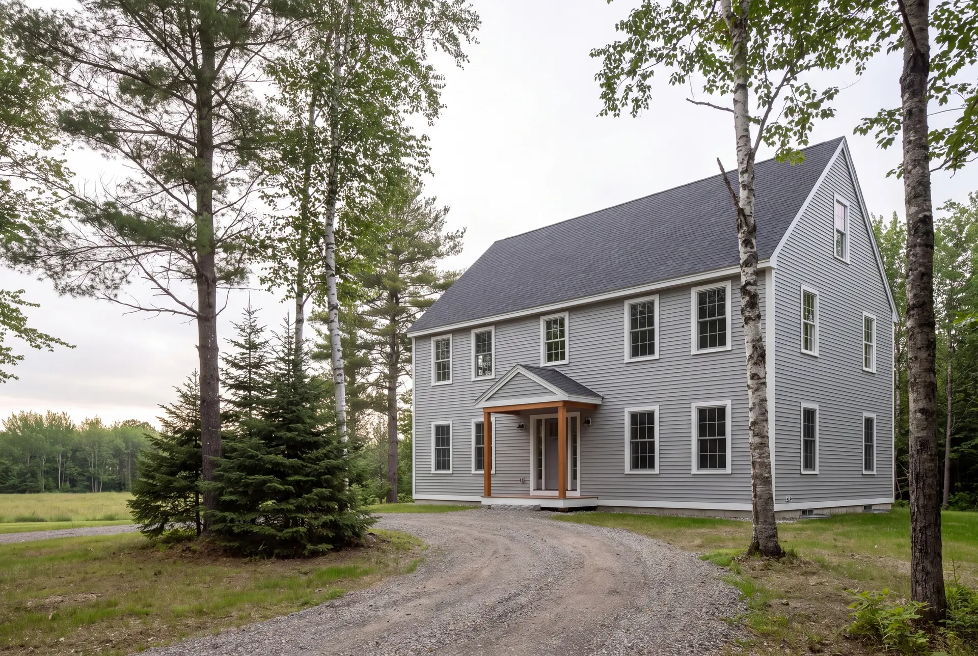 Illustration: A newly built gray saltbox house with a dark roof and white windows, seen from a gravel driveway surrounded by trees