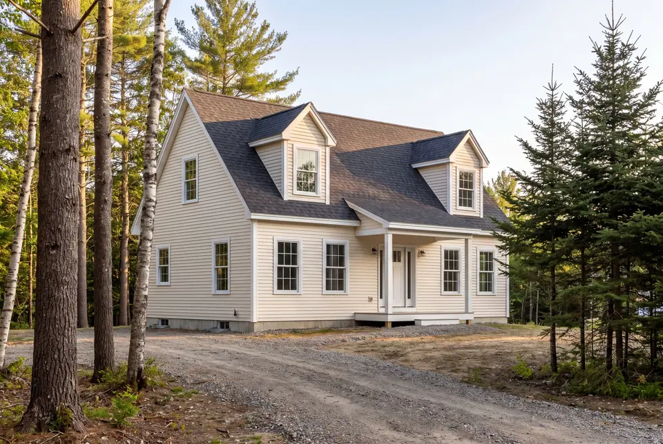 Illustration: A newly built Cape house with cream siding and a dark roof, nestled among tall trees, with a gravel driveway leading to the front
