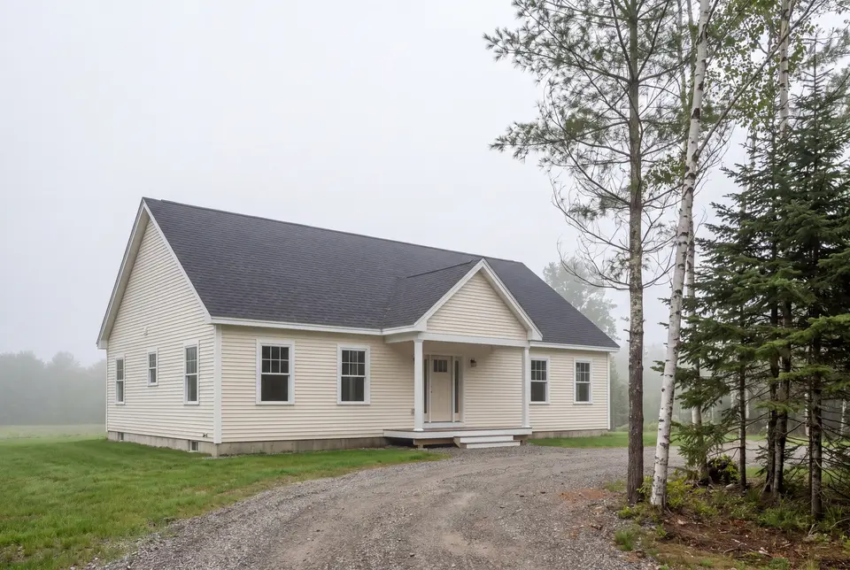 general contractor in Windham, ME: illustrative scene, A newly built, light cream colored ranch house with a dark gray roof, surrounded by green grass and a gravel driveway, with trees on the right and a misty background
