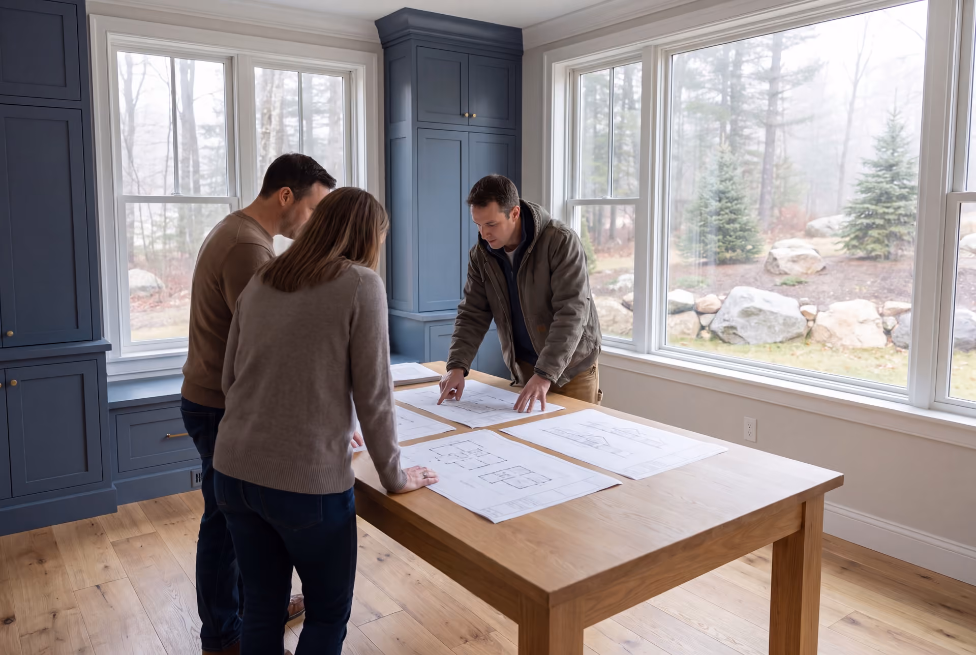house plans and design in Old Orchard Beach, ME: illustrative scene, Three people, a builder and two homeowners, reviewing architectural drawings on a table in a finished living room with blue cabinets and oak floors