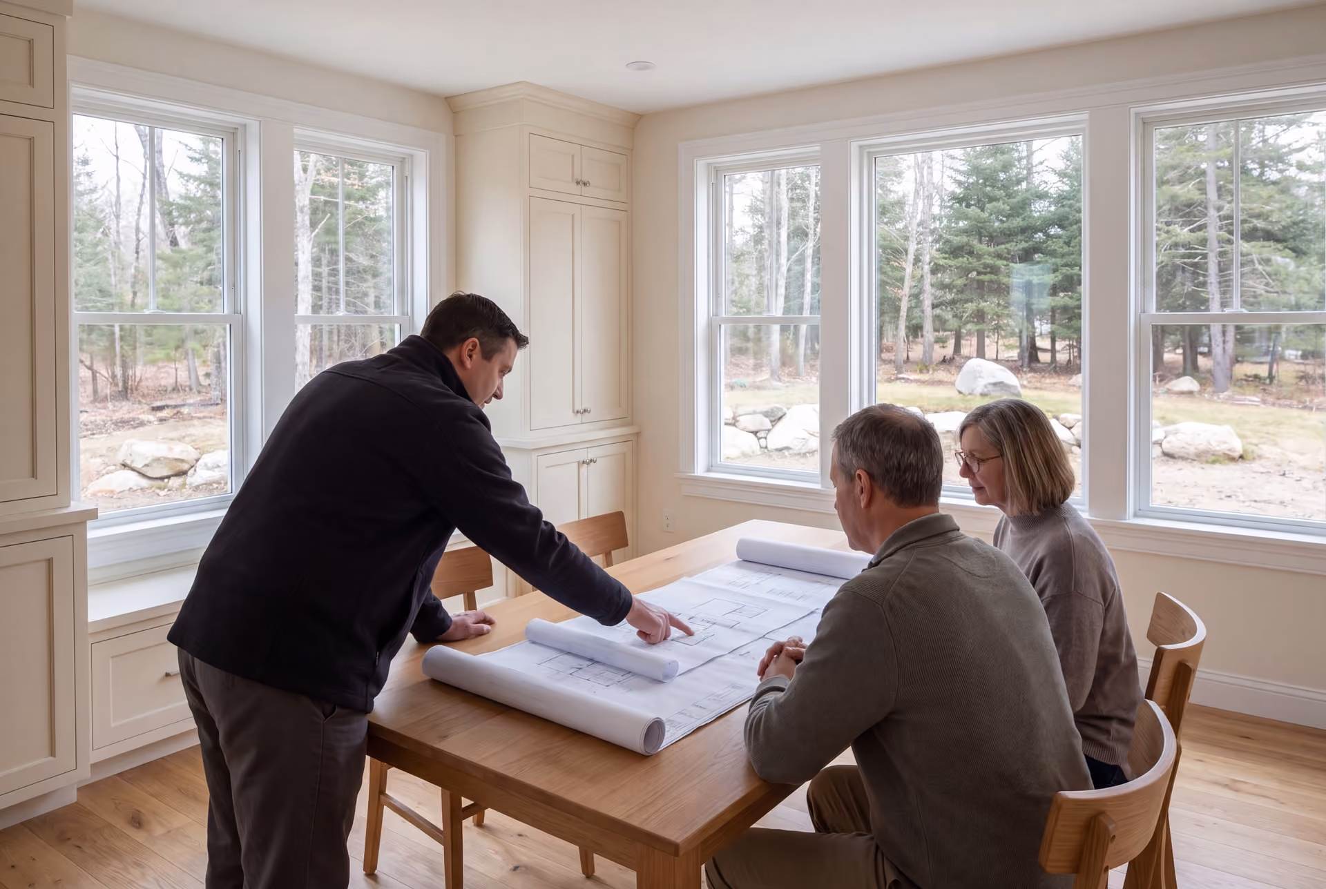 house plans and design in South Portland, ME: illustrative scene, Three adults, a builder and two homeowners, reviewing architectural drawings on an oak table in a finished living room
