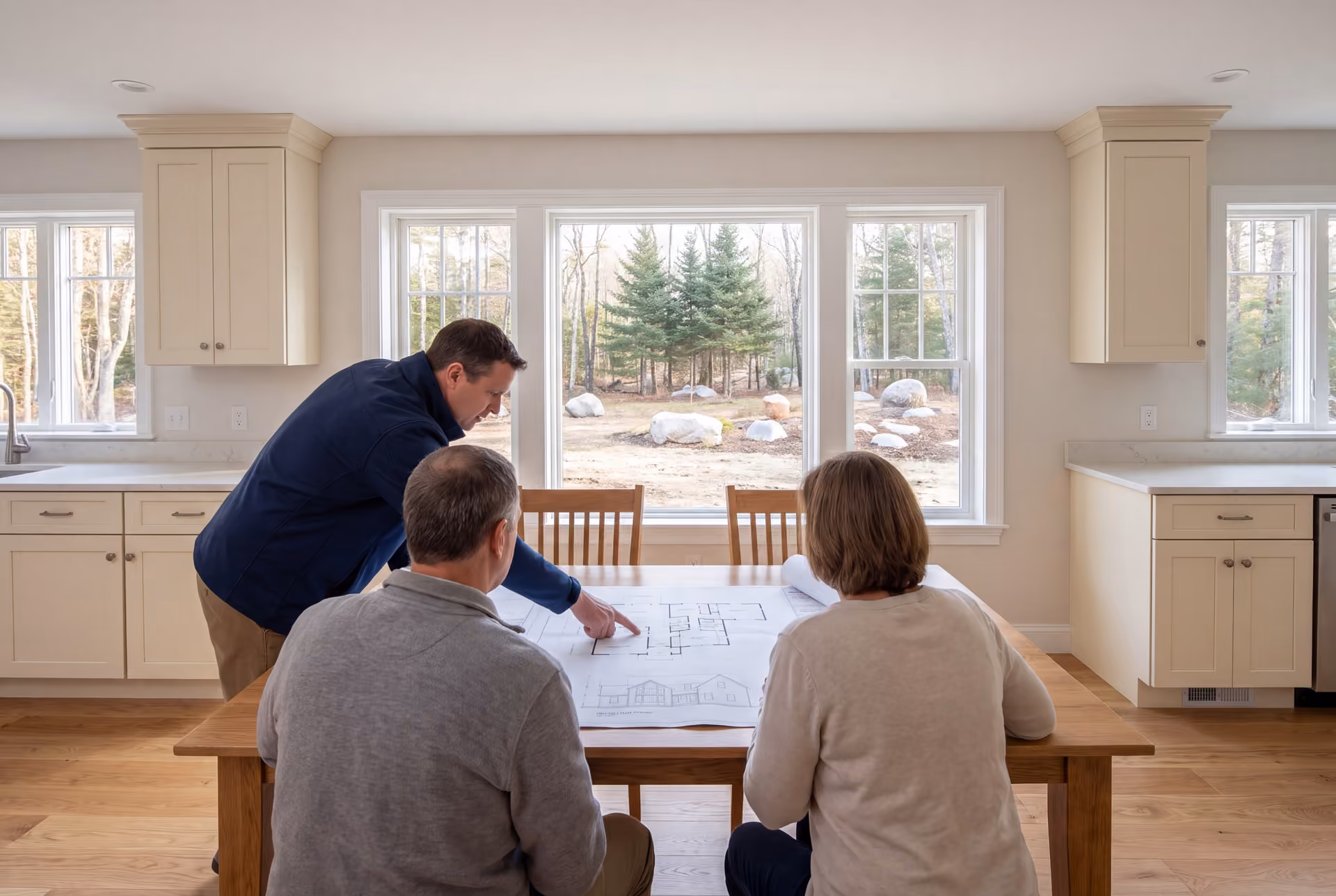 house plans and design in Standish, ME: illustrative scene, Three people, a builder and two homeowners, reviewing architectural drawings on an oak table in a finished living room with cream cabinetry and oak floors